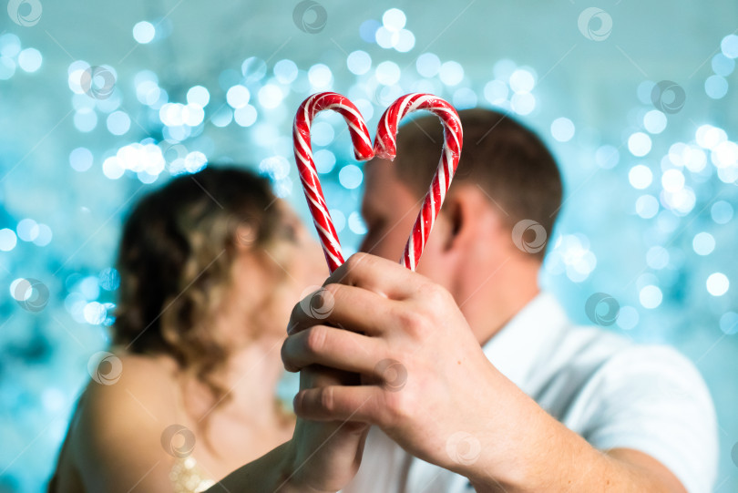 Скачать влюбленная пара целуется и сердечко сделано из двух леденцов на палочке. день святого Валентина. В руках держите две леденцовые палочки в форме сердца. Сердечко, сформированное из двух белых и красных рождественских конфет. фотосток Ozero