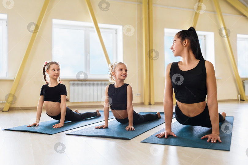 Скачать Женщина-тренер и две маленькие девочки, практикующие йогу в студии, выполняют упражнение с собакой мордой вверх на гимнастических ковриках, поза Урдхва Мукха Шванасана, групповая тренировка фотосток Ozero