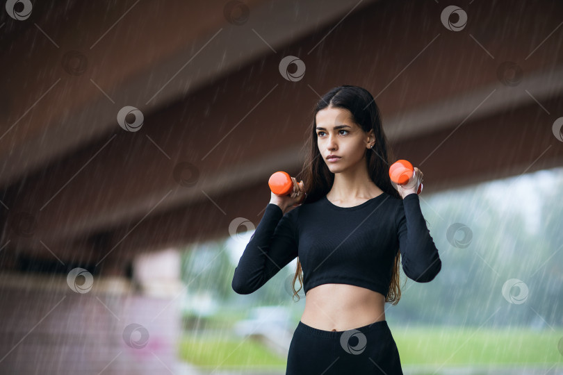 Скачать Красивая молодая девушка выполняет упражнения с гантелями во время дождя, концепция здорового образа жизни фотосток Ozero