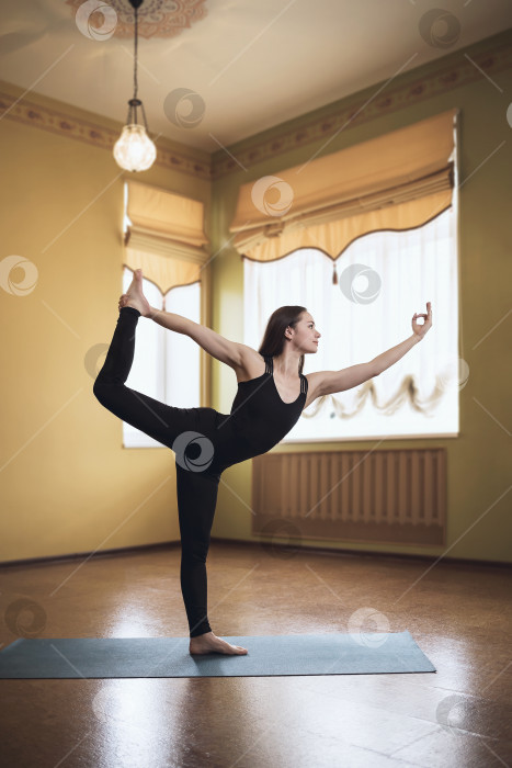 Скачать Женщина в черной спортивной одежде, практикующая йогу, выполняет упражнение натараджасана, позу короля танцоров. Модель тренируется в комнате на коврике фотосток Ozero