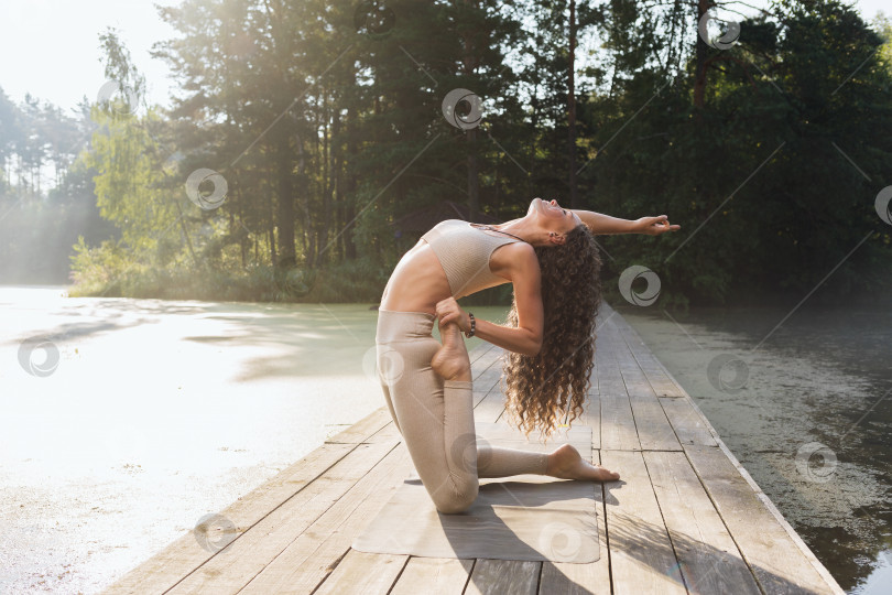 Скачать Женщина в спортивной одежде, практикующая йогу, выполняющая упражнение Уштрасана с вирасаной, позой верблюда и героя, тренирующаяся на открытом воздухе в парке солнечным утром у пруда фотосток Ozero