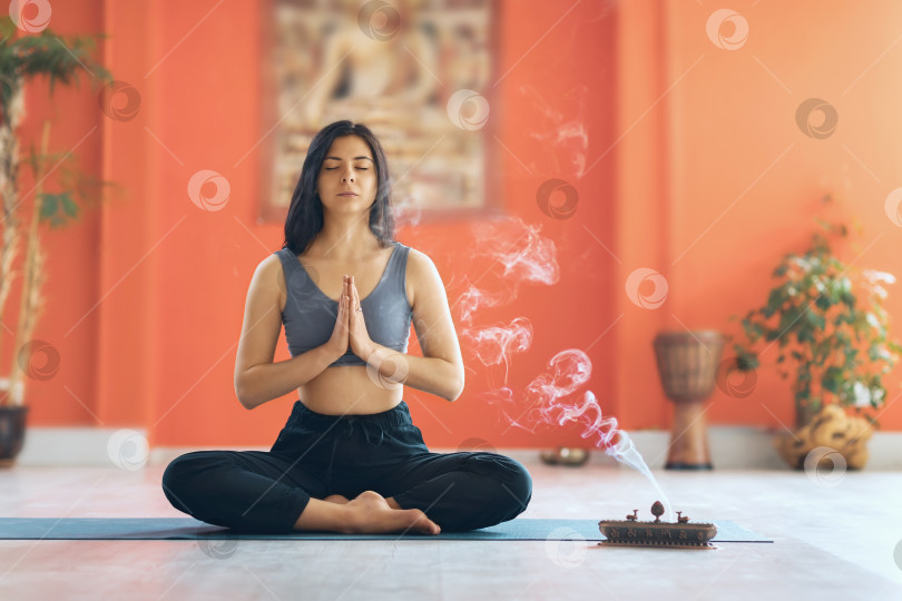 Скачать Молодая женщина в спортивной одежде, практикующая йогу, сидя на коврике в позе лотоса, занимается медитацией с намасте в студии фотосток Ozero