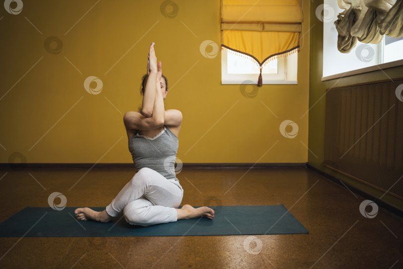 Скачать Женщина в серой спортивной одежде, практикующая йогу, выполняющая упражнение гомукхасана, поза коровы, тренирующаяся сидя на коврике в студии фотосток Ozero