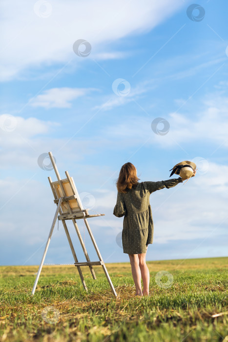 Скачать Вдохновленная молодая женщина-художница в платье стоит в поле с мольбертом теплым солнечным вечером фотосток Ozero
