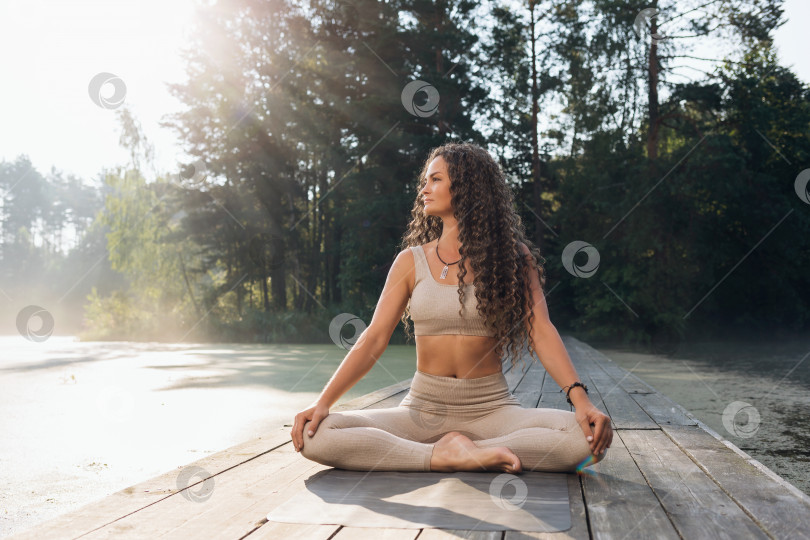Скачать Красивая женщина в бежевой спортивной одежде, практикующая йогу, сидя на коврике в позе лотоса, занимается медитацией на берегу пруда теплым солнечным утром фотосток Ozero