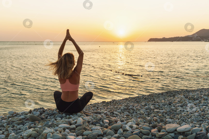 Скачать Женщина в спортивной одежде, практикующая йогу, медитирует с поднятыми вверх руками, сидя в позе лотоса на галечном морском берегу летним солнечным вечером фотосток Ozero