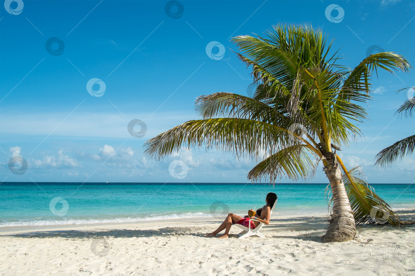 Скачать Молодая женщина отдыхает в кресле под пальмой на берегу Индийского океана, глядя вдаль фотосток Ozero