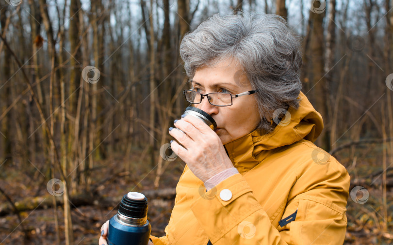 Скачать Пожилая женщина в очках, пьющая горячий чай, крупным планом. Пожилая женщина гуляет по лесу. Концепция согревающего напитка в холодную погоду, активного образа жизни на пенсии. фотосток Ozero