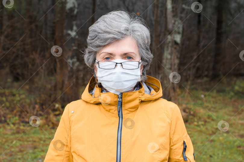 Скачать Портрет пожилой женщины в очках и медицинской маске для лица. Пожилая женщина в защитной маске смотрит в камеру снаружи. фотосток Ozero
