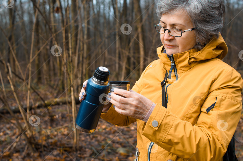 Скачать Пожилая женщина в очках прогуливается по лесу. Пожилая женщина держит походный термос с горячим напитком. Концепция согревающего напитка в холодную погоду, активного образа жизни на пенсии. фотосток Ozero