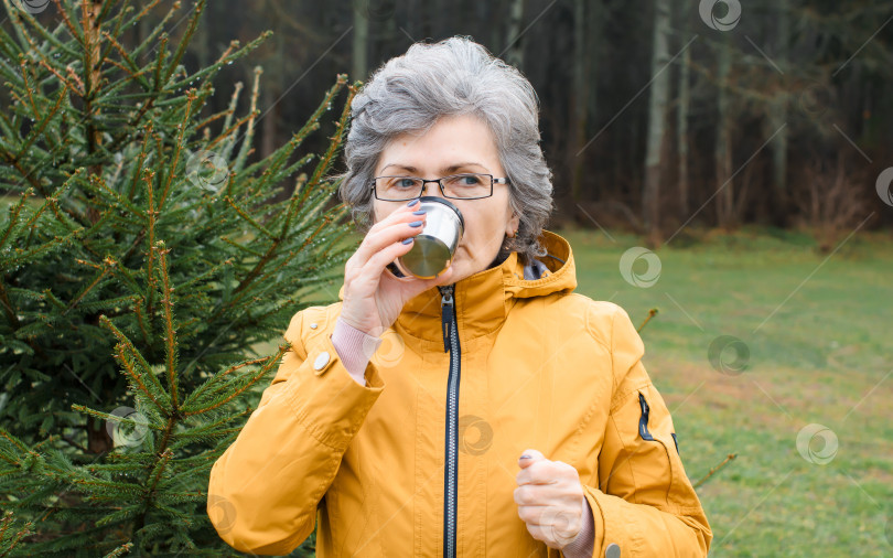 Скачать Пожилая женщина пьет горячий чай в лесу на открытом воздухе. Портрет пожилой женщины, прогуливающейся в холодную погоду. фотосток Ozero