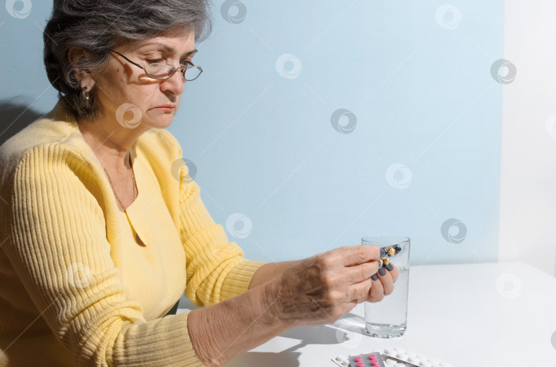 Скачать Пожилая женщина в очках читает инструкцию к лекарству. Пенсионерка держит дома таблетки и стакан воды. Концепция лечения, выход на пенсию по болезни фотосток Ozero
