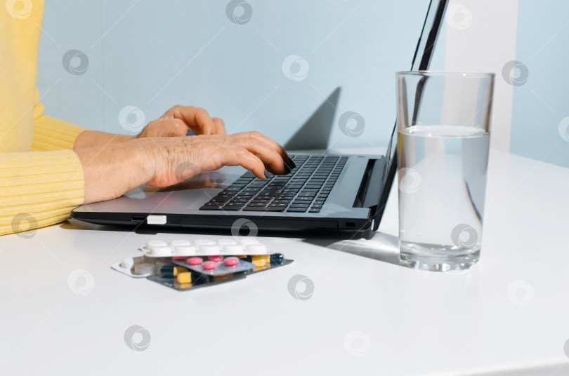 Скачать Руки пожилой женщины печатают на ноутбуке крупным планом. Лекарства и стакан воды стоят дома на столе. Концепция онлайн-консультации с врачом, поиск инструкций в Интернете фотосток Ozero