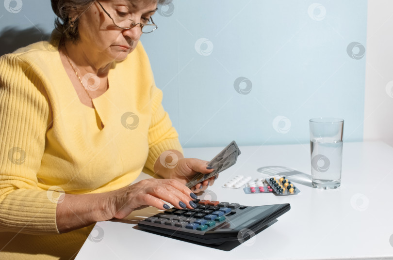 Скачать Пожилая женщина держит в руках деньги и считает на калькуляторе. Пенсионерка сидит на кухне с лекарствами. Понятие депрессии, цена на лекарства, стоимость лечения фотосток Ozero