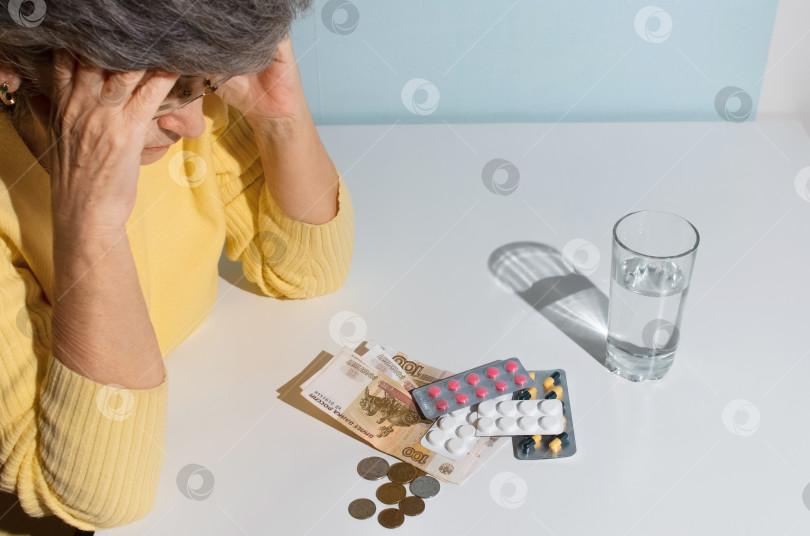 Скачать Пожилая женщина в очках смотрит на деньги и таблетки на столе. Понятие депрессии, цена на лекарства, стоимость лечения фотосток Ozero