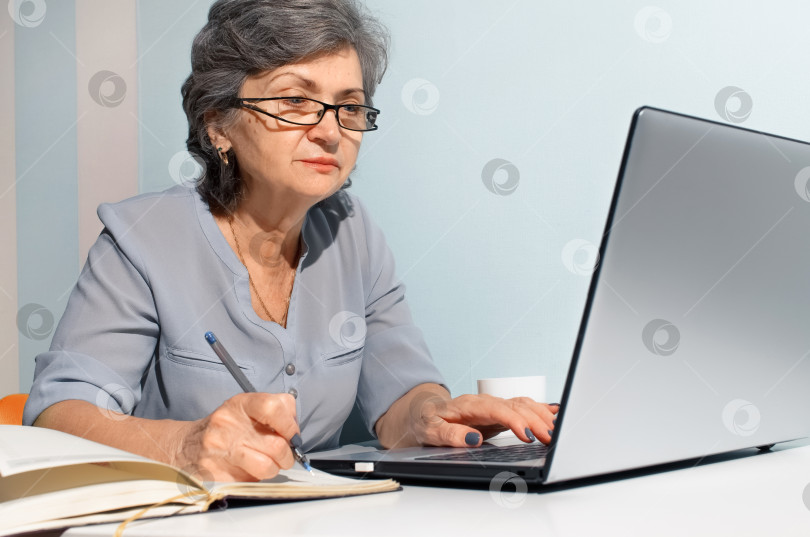 Скачать Пожилая женщина пользуется ноутбуком и пишет в дневнике. Концепция удаленной работы, фриланса, работы на пенсии фотосток Ozero