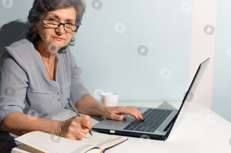 Скачать Пожилая женщина пользуется ноутбуком и пишет в дневнике. Пожилая женщина-портье смотрит в камеру. Концепция удаленной работы, фриланса, работы на пенсии фотосток Ozero
