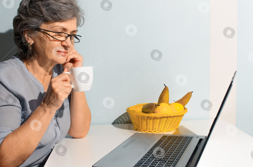 Скачать Пожилая женщина пользуется ноутбуком дома. Пожилая женщина пьет кофе и смотрит на экран ноутбука. Концепция видеозвонка, новая норма, самоизоляция фотосток Ozero