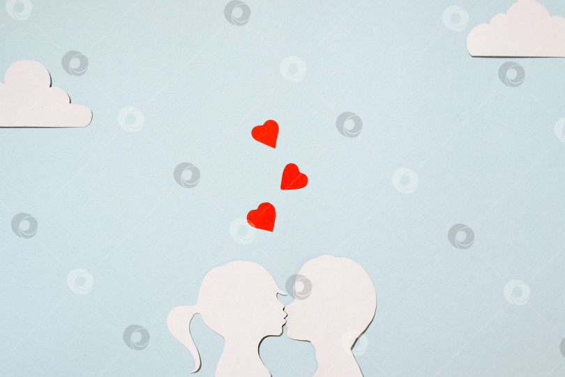 Скачать Концепция Дня святого Валентина. Картонные силуэты целующихся девочки и мальчика. Креативная любовная открытка. Красные сердечки, облака на пастельно-голубом фоне. Ровное положение, вид сверху. фотосток Ozero