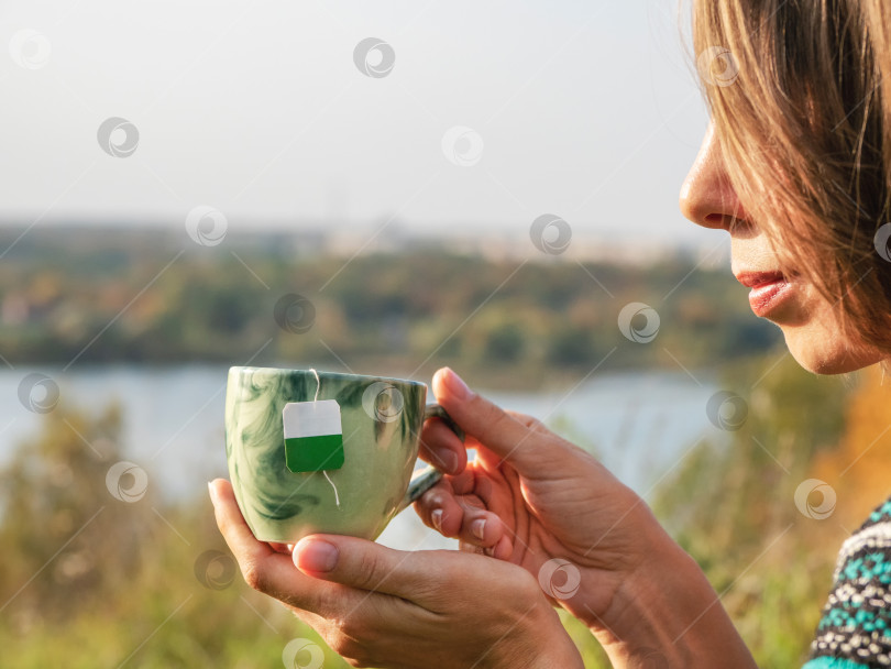 Скачать Женские руки держат фарфоровую кружку с пакетиком внутри, пьют горячий зеленый чай на природе. Женщина наслаждается теплым варевом или напитком в чашке, расслабляется, отдыхая во время перерыва на свежем воздухе фотосток Ozero