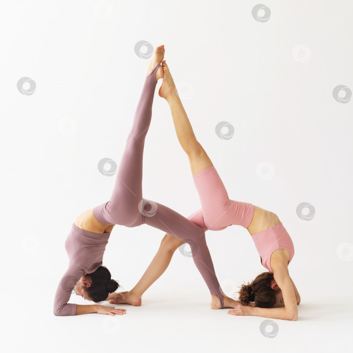 Скачать Две женщины, практикующие йогу, выполняющие парное упражнение Эка Пада Випарита Дандасана, поза перевернутого посоха, тренируются в студии на ярком фоне фотосток Ozero