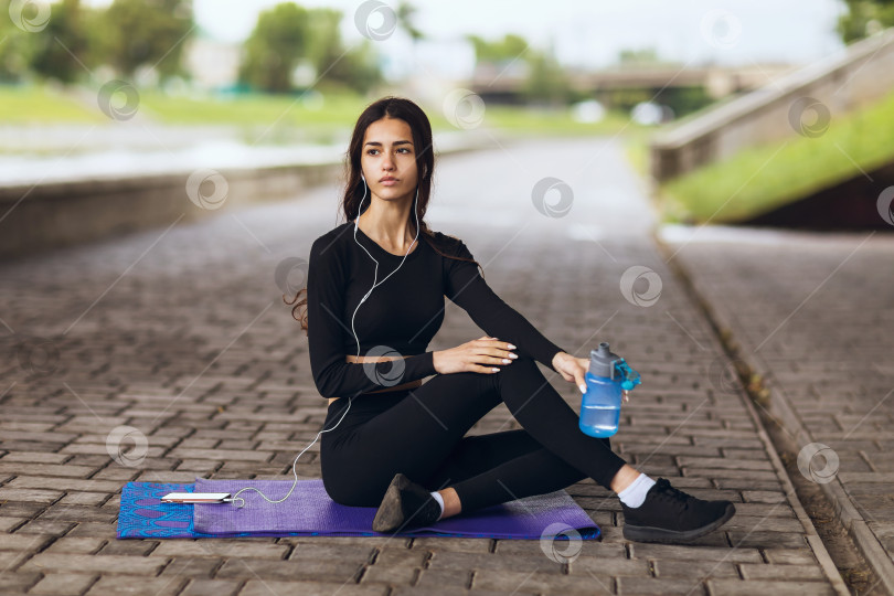 Скачать Молодая женщина сидит на гимнастическом коврике на набережной и отдыхает после занятий спортом фотосток Ozero