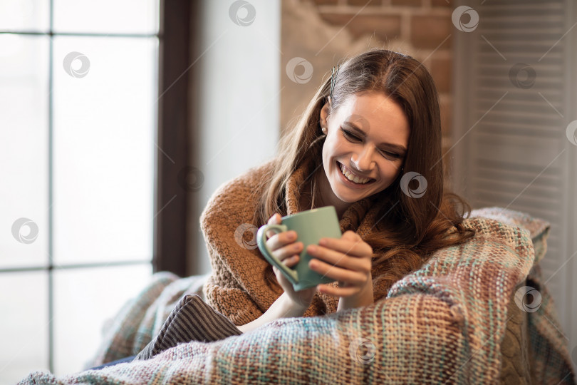 Скачать Фотография в домашней обстановке молодой красивой девушки, сидящей в кресле с кружкой кофе и улыбающейся от положительных эмоций фотосток Ozero