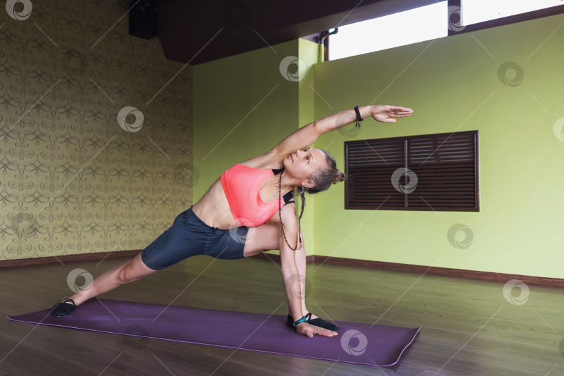 Скачать Женщина, практикующая йогу, выполняет упражнение уттхита паршваконасана, позу угла с интенсивным боковым растяжением, стоит на коврике в студии фотосток Ozero