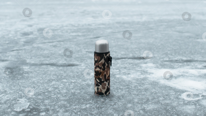 Скачать Термос в футляре цвета хаки, стоящий на замерзшем озере. Концепция горячего напитка в холодную погоду, на зимней рыбалке, в походе фотосток Ozero