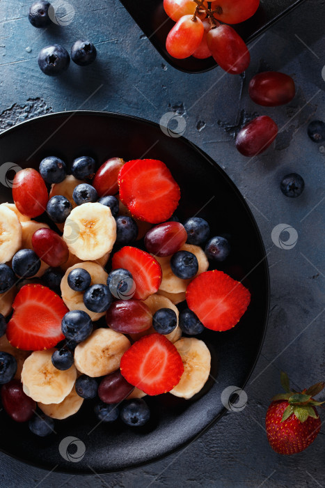 Скачать Полезный завтрак из фруктов и ягод, вид сверху. Нарезанные бананы, клубнику, виноград и чернику выложить на черную тарелку фотосток Ozero