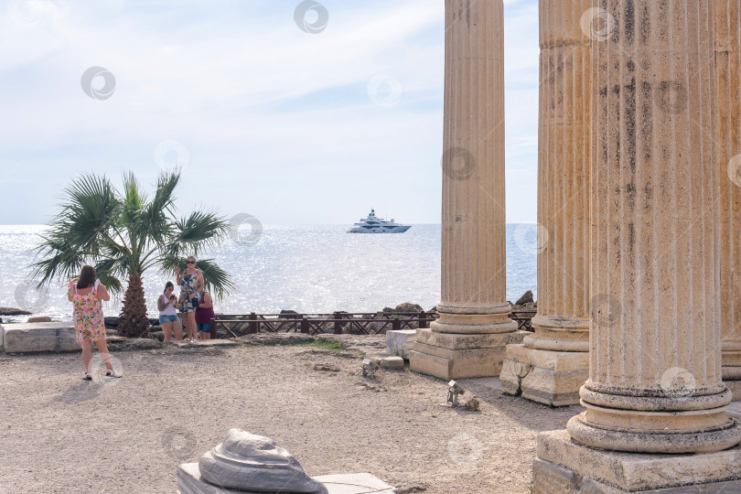 Скачать туристы фотографируются под пальмой на берегу моря среди древних руин в Сиде, Турция фотосток Ozero