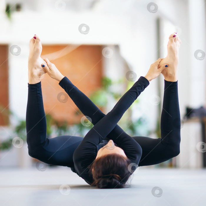 Скачать Женщина в черной спортивной одежде, практикующая йогу, выполняет упражнение супта рудрасана, тренируясь лежа на коврике в комнате фотосток Ozero