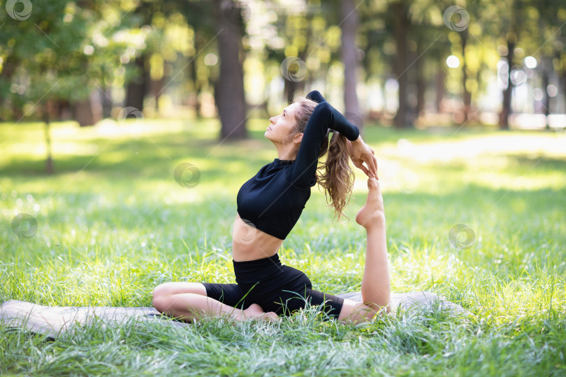 Скачать Молодая женщина в черной спортивной одежде, занимающаяся йогой, выполняет вариацию упражнения Эка Пада Раджакапотасана, поза голубя, тренируется в парке теплым летним утром, сидя на коврике фотосток Ozero