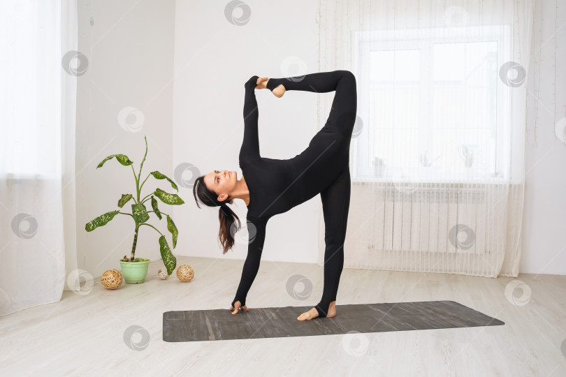 Скачать Женщина в черной спортивной одежде, практикующая йогу, выполняет вариацию упражнения Ардха Чандрасана, позу полумесяца, тренируясь, стоя на коврике в комнате у окна фотосток Ozero