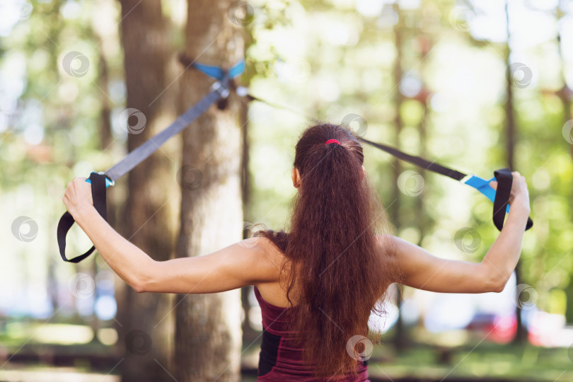 Скачать Молодая женщина выполняет упражнение для проработки мышц спины на подвесном тренажере, прикрепленном к дереву в парке фотосток Ozero