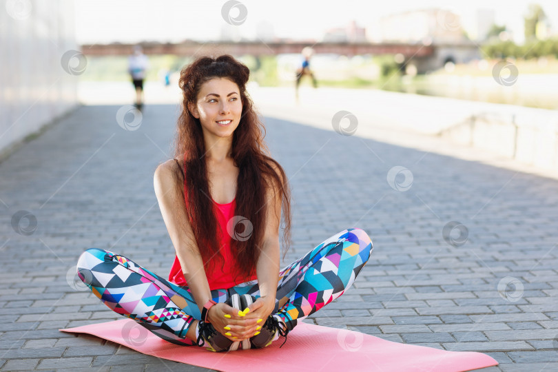Скачать Молодая женщина выполняет утреннюю зарядку, сидя на гимнастическом коврике на открытом воздухе фотосток Ozero