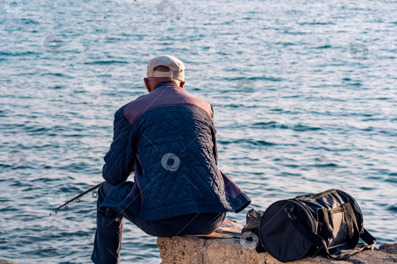 Скачать пожилой рыбак сидит с удочкой на прибрежном камне фотосток Ozero