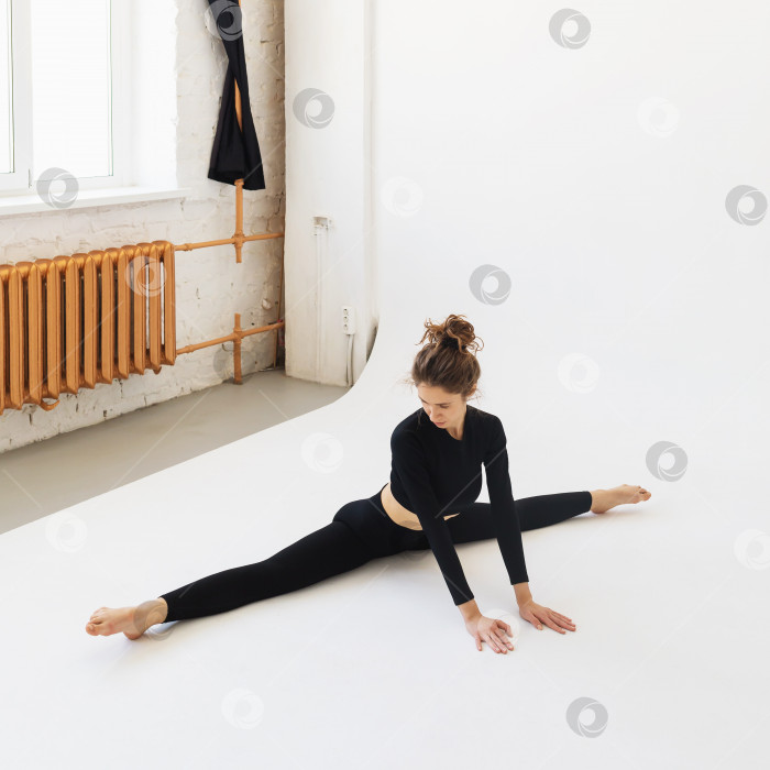 Скачать Молодая женщина в черной спортивной одежде, занимается растяжкой мышц ног, выполняет поперечный шпагат, сидя на белом фоне в студии фотосток Ozero