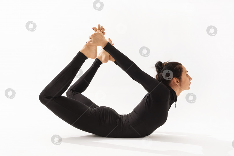 Скачать Женщина в черной спортивной одежде, практикующая йогу, выполняет упражнение дханурасана, поза лука, на белом фоне фотосток Ozero