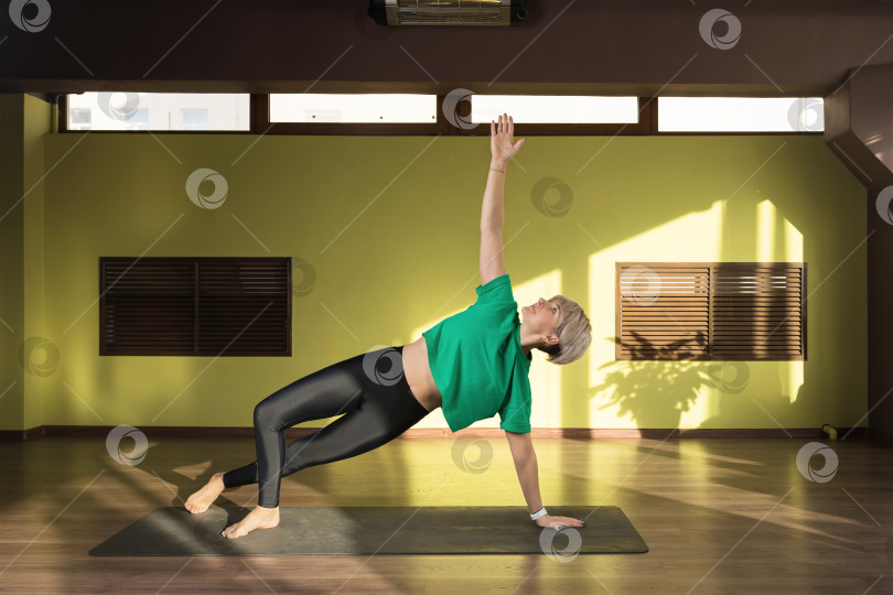 Скачать Женщина, практикующая йогу, выполняющая вариацию упражнения васиштхасана, позу боковой доски, стоя на коврике в студии фотосток Ozero