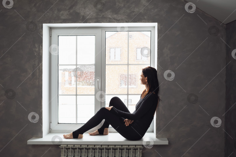 Скачать Женщина в спортивной одежде сидит на подоконнике и смотрит в окно на снегопад фотосток Ozero