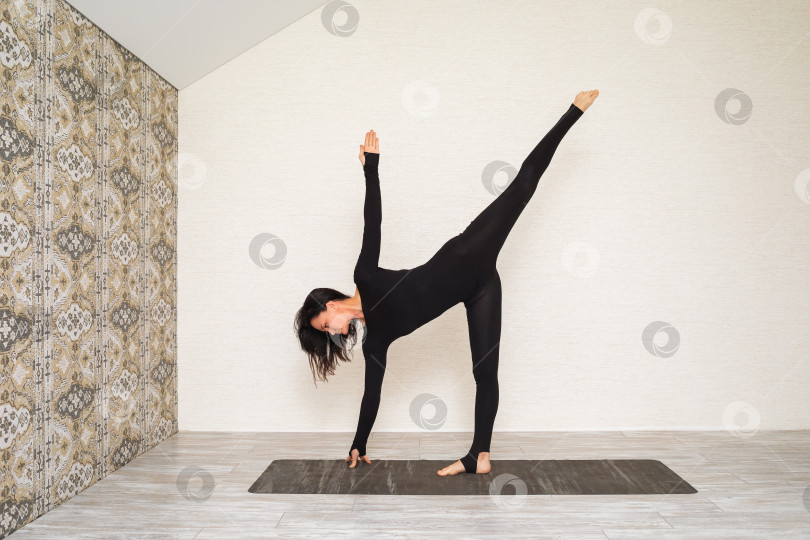 Скачать Женщина в черной спортивной одежде, практикующая йогу, выполняет упражнение Ардха Чандрасана, поза полумесяца, тренируется, стоя на коврике в комнате у стены фотосток Ozero