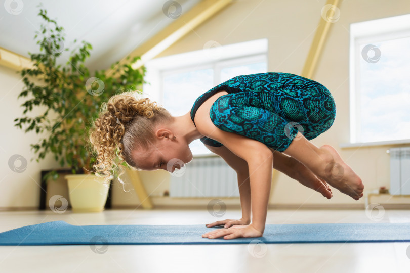 Скачать Маленькая красивая девочка со светлыми волосами практикует йогу, выполняя упражнение "стойка на руках", какасану или позу вороны на гимнастическом коврике в студии фотосток Ozero