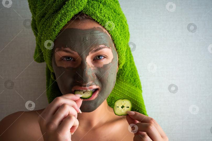 Скачать Красивая молодая женщина с маской для лица на лице, держащая ломтики огурца. фотосток Ozero