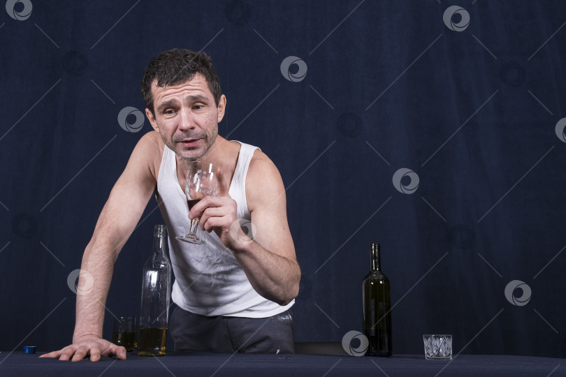 Скачать Пьяный одинокий мужчина в повседневной домашней одежде стоит за столом на темном фоне. На столе стоят бутылки со спиртными напитками. В руке держит фужер с вином. И старается что-то сказать. Алкоголизм как болезнь человечества. фотосток Ozero