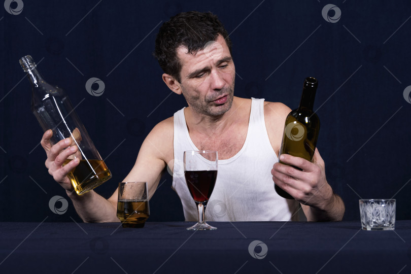 Скачать Пьяный небритый мужчина средних лет в домашней белой майке сидит в комнате на темном фоне. Перед ним стоят фужер и бокал, наполненные алкогольными напитками. В своих руках он держит открытые бутылки с алкоголем и смотрит на них. Алкоголизм как болезнь человечества. фотосток Ozero