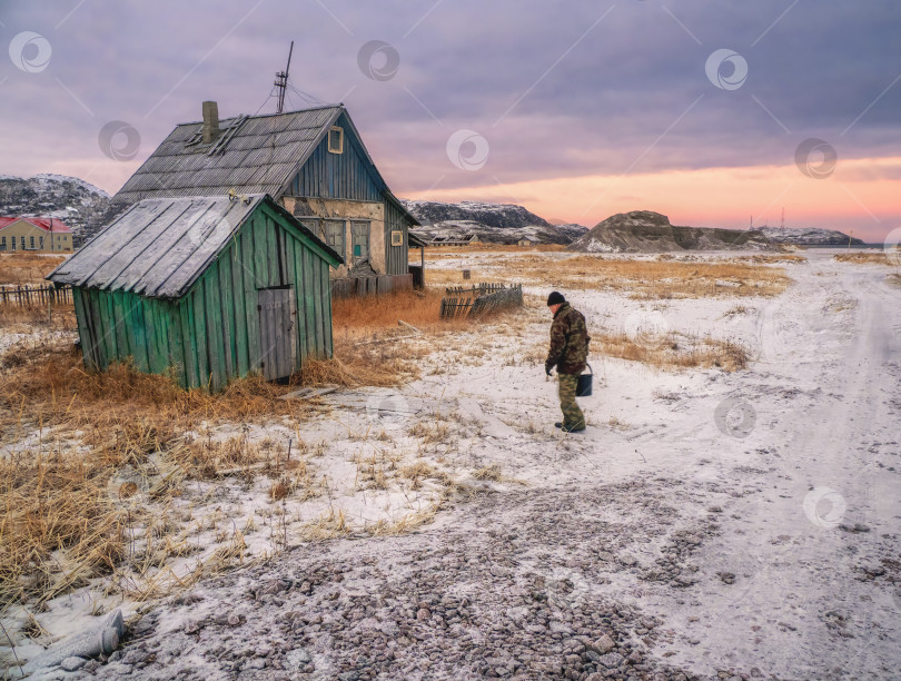 Скачать Аутентичная русская северная деревня, старые полуразрушенные деревянные дома, суровая арктическая природа. Мужчина с ведром воды идет к себе домой. фотосток Ozero