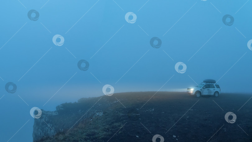 Скачать Мягкий фокус. Плохая видимость в непогоду. Белый туристический автомобиль в ночном тумане с включенными фарами на краю обрыва. Экстремальное горное сафари. фотосток Ozero