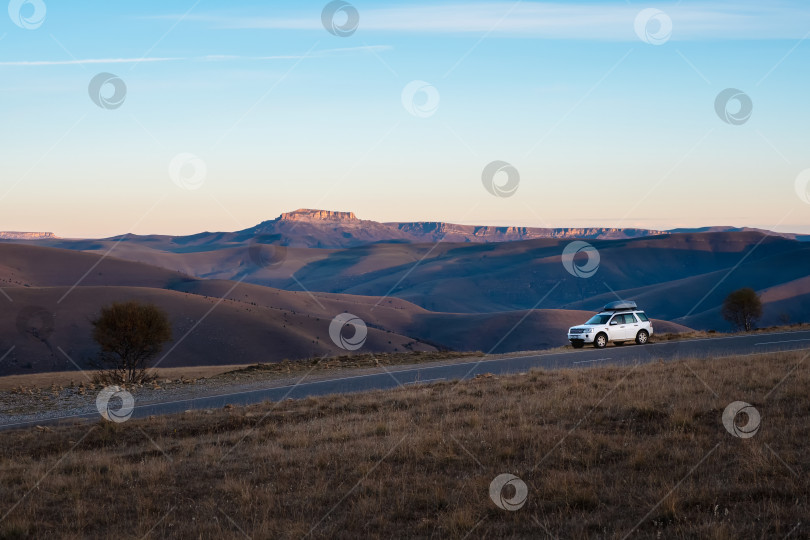 Скачать Белый автомобиль-внедорожник припаркован рядом с живописной трассовой дорогой в утреннем осеннем пейзаже недалеко от плато Бермамыт. Гора багажных ящиков на багажнике на крыше автомобиля. фотосток Ozero