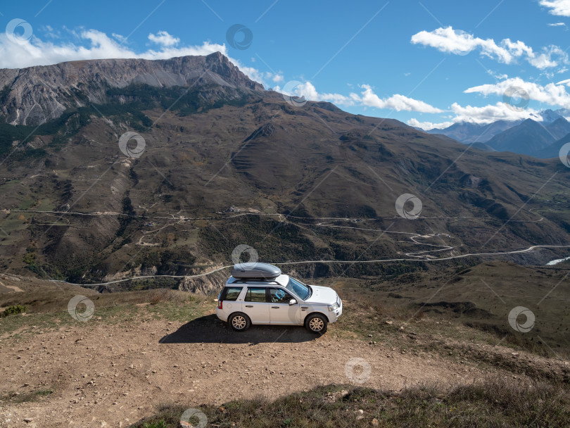 Скачать Белый внедорожник Rover car едет по узкой горной дороге на фоне острых горных скал. Гора багажных ящиков на багажнике на крыше автомобиля. фотосток Ozero